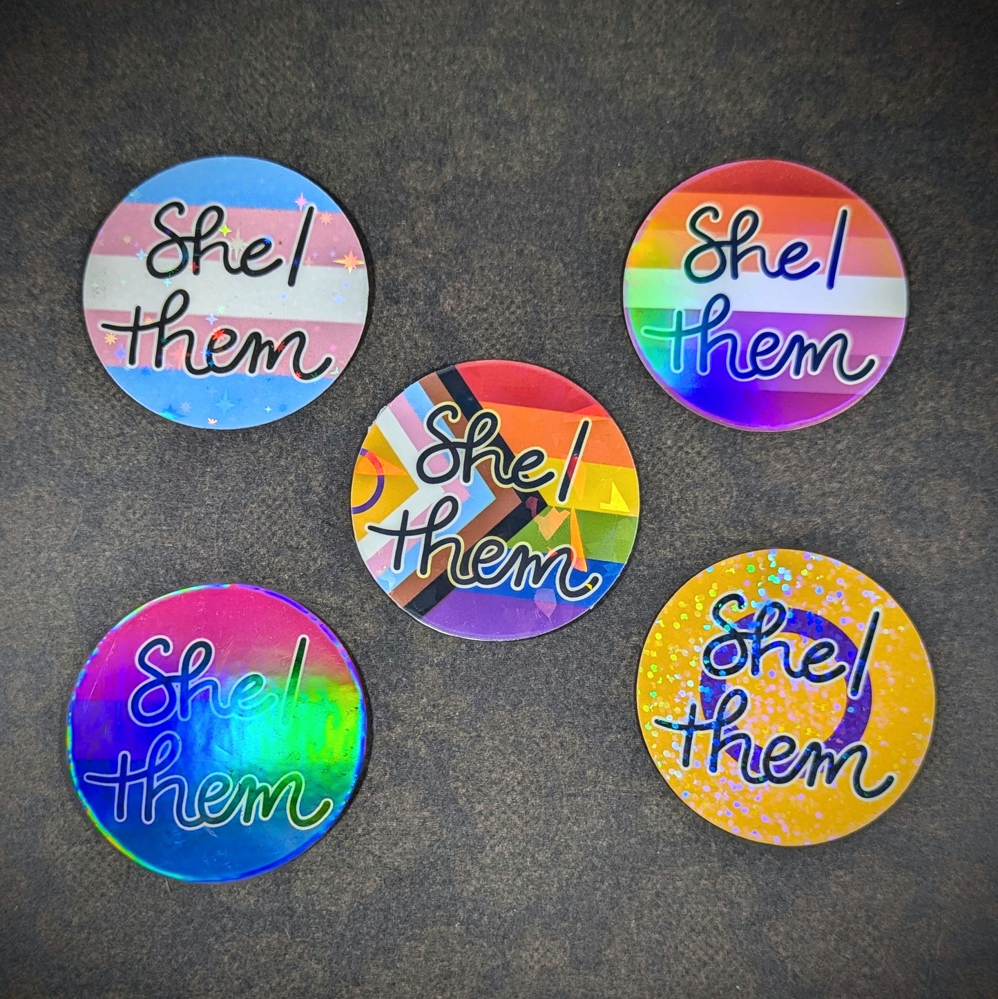 "Round holographic pronoun stickers featuring 'She them' in various LGBTQ+ pride flag designs, perfect for expressing gender identity and inclusivity on laptops, water bottles, and notebooks."