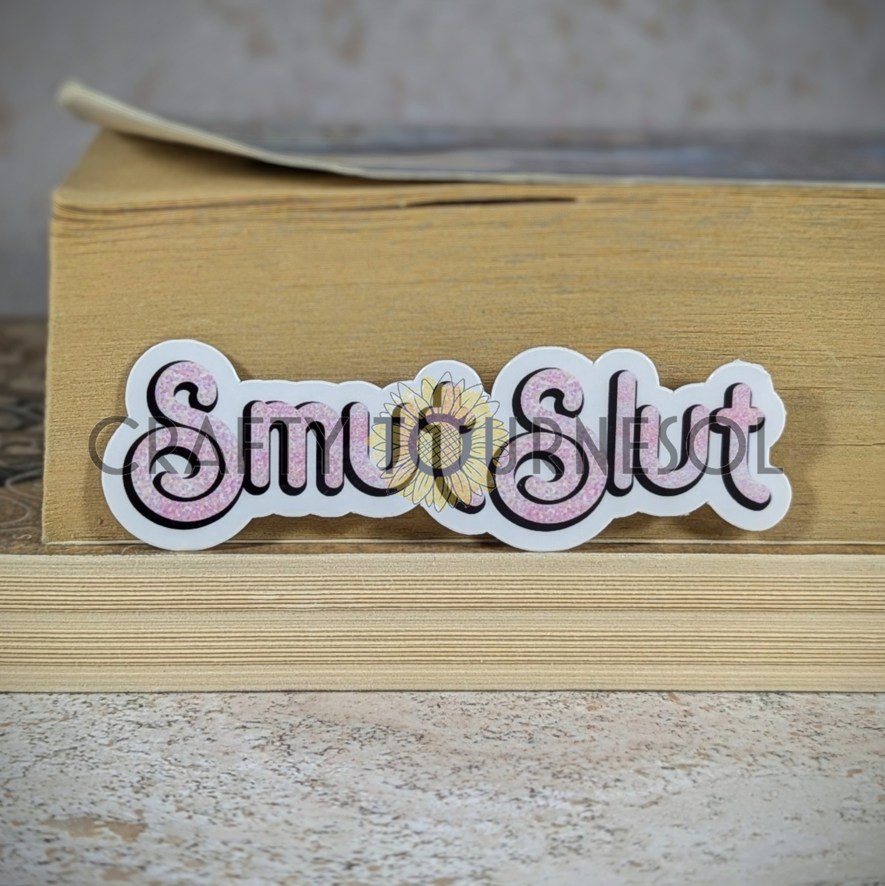 "Smut Slut" sticker with elegant pink speckled text, perfect for book lovers and spice enthusiasts. Set against a cozy book stack background, this design adds a bold, fun flair to your aesthetic.