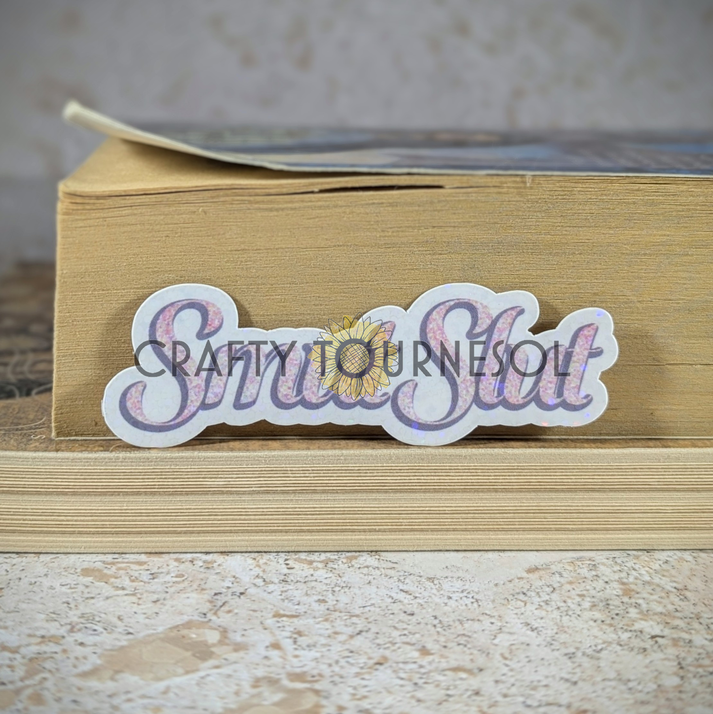 "Smut Slut" holographic sticker with elegant pink speckled text, perfect for book lovers and spice enthusiasts. Set against a cozy book stack background, this design adds a bold, fun flair to your aesthetic.