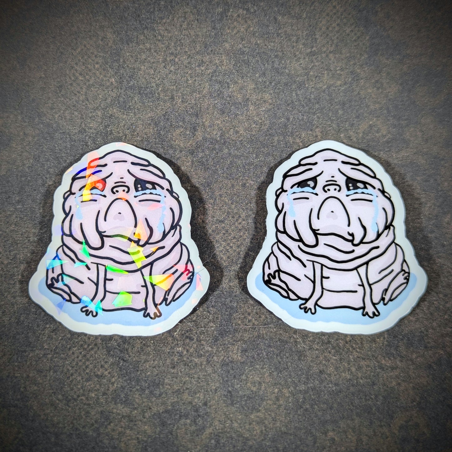 "Pair of Squonk stickers featuring a sad, mythical creature design, with one holographic and one matte version. Perfect for cryptid enthusiasts and collectors of unique, emotional supernatural decor."