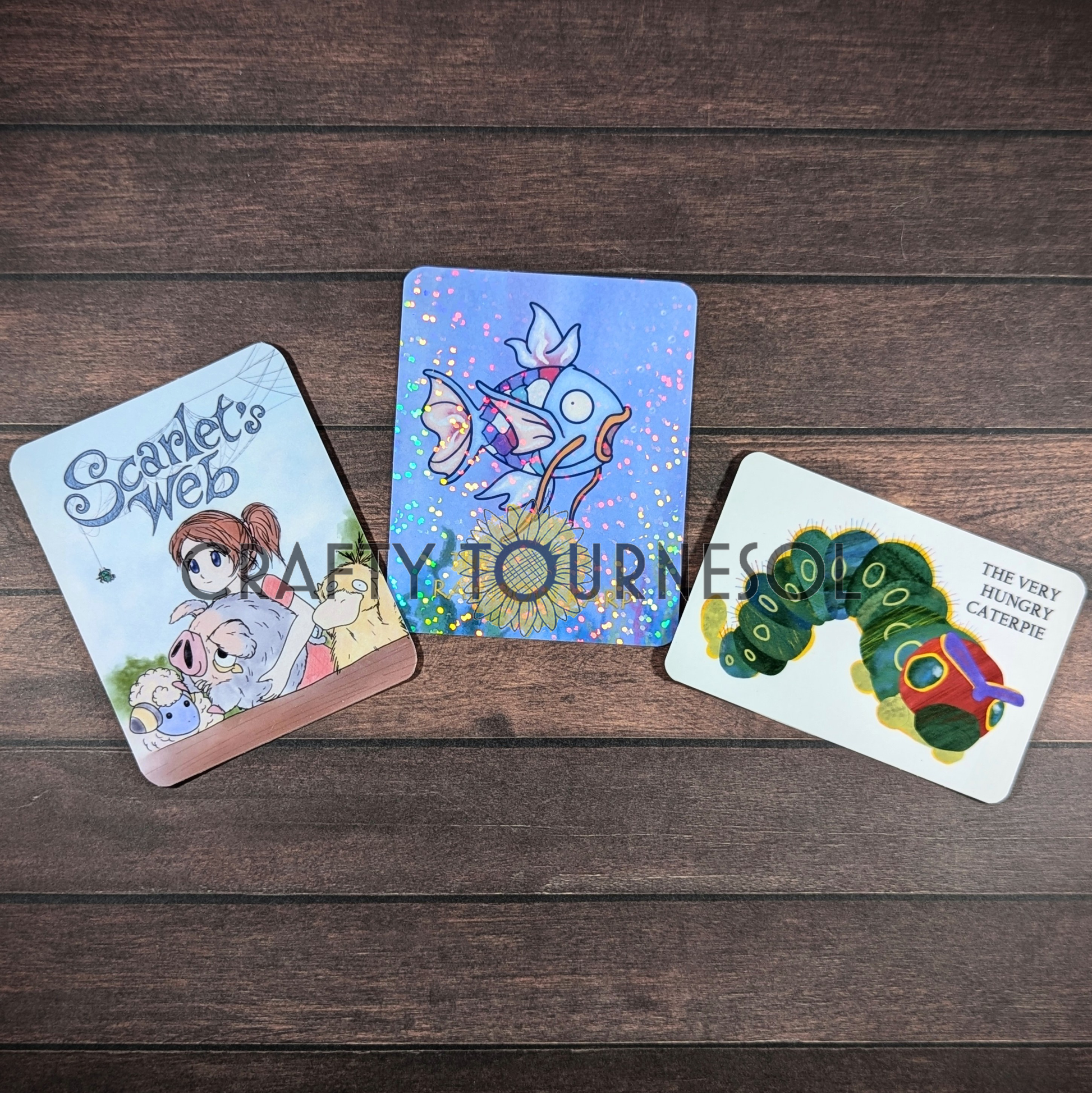 
"Vinyl stickers inspired by storybook themes: Scarlet's Web, The Rainbow Karp, and The Very Hungry Caterpie, designed for collectors and gamers, displayed on a wooden background."