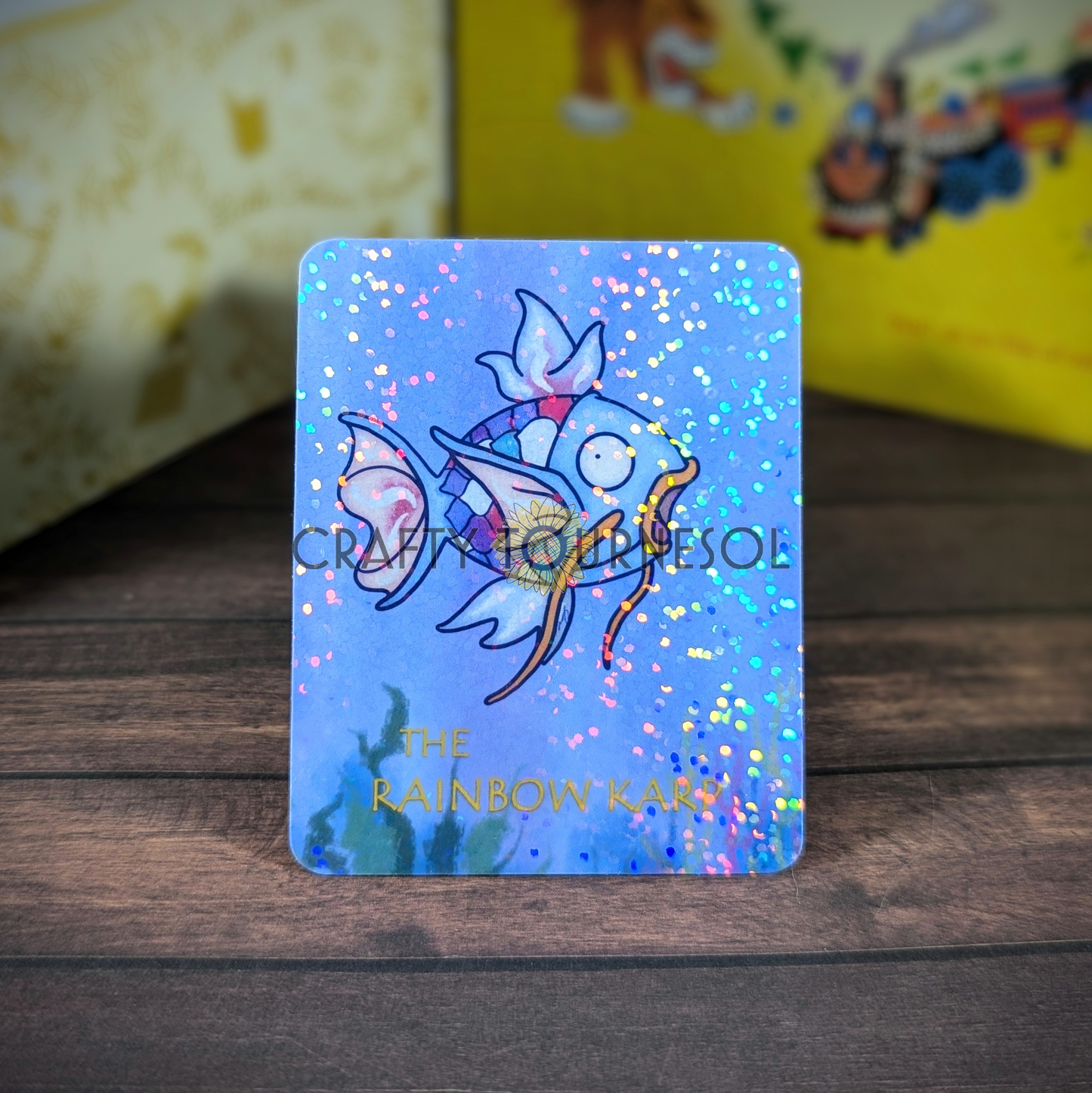 "Vinyl sticker titled The Rainbow Karp, featuring a blue watercolor Magikarp styled like the children's book character The Rainbow Fish, with holographic finish, blending storybook charm and gaming aesthetics, set against a soft wooden and blurred background."