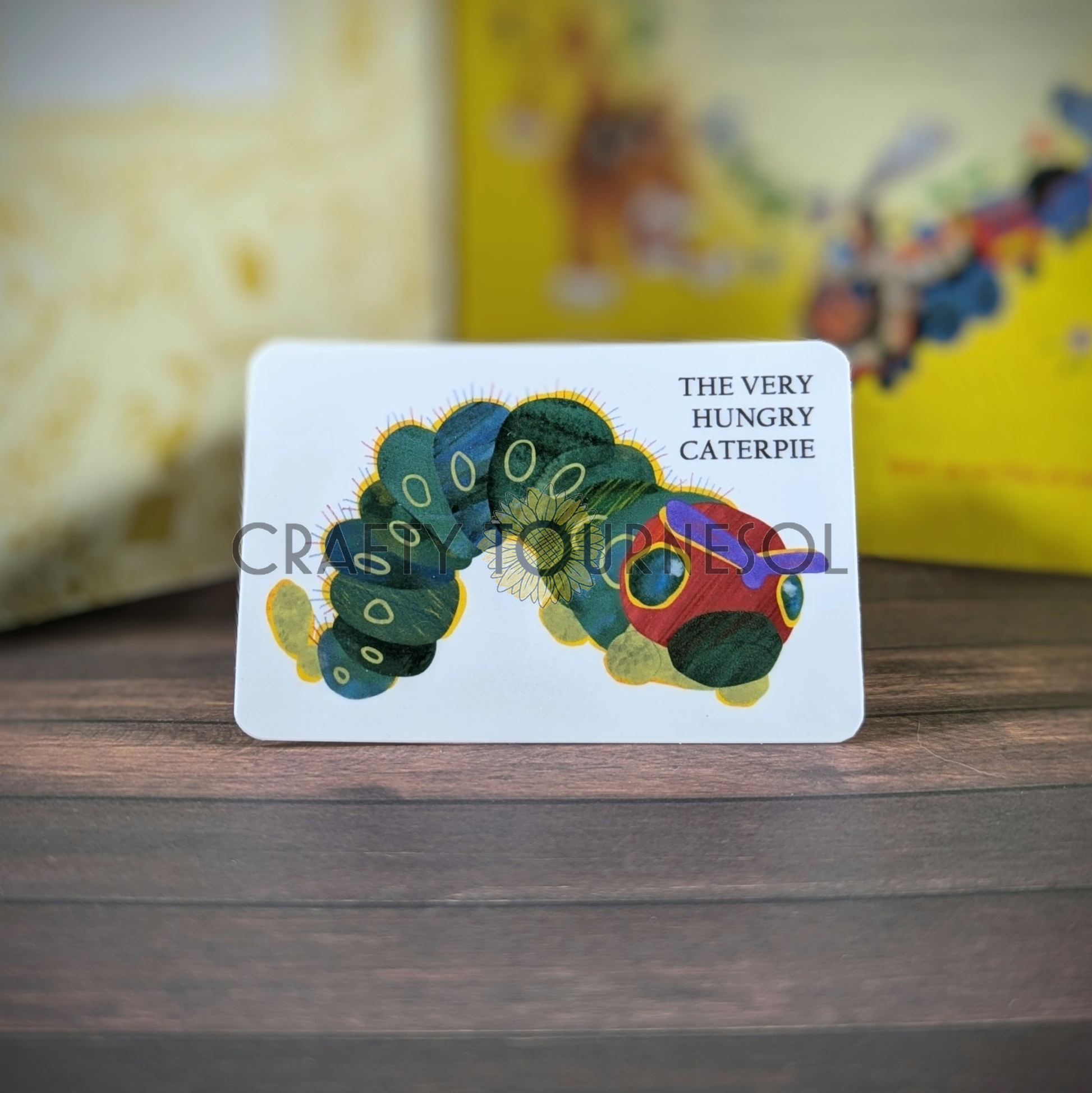 "Vinyl sticker titled The Very Hungry Caterpie, with a Caterpie drawn and colored like the children's storybook character The Very Hungry Caterpillar, blending storybook charm and gaming aesthetics, set against a soft wooden, blurred background."