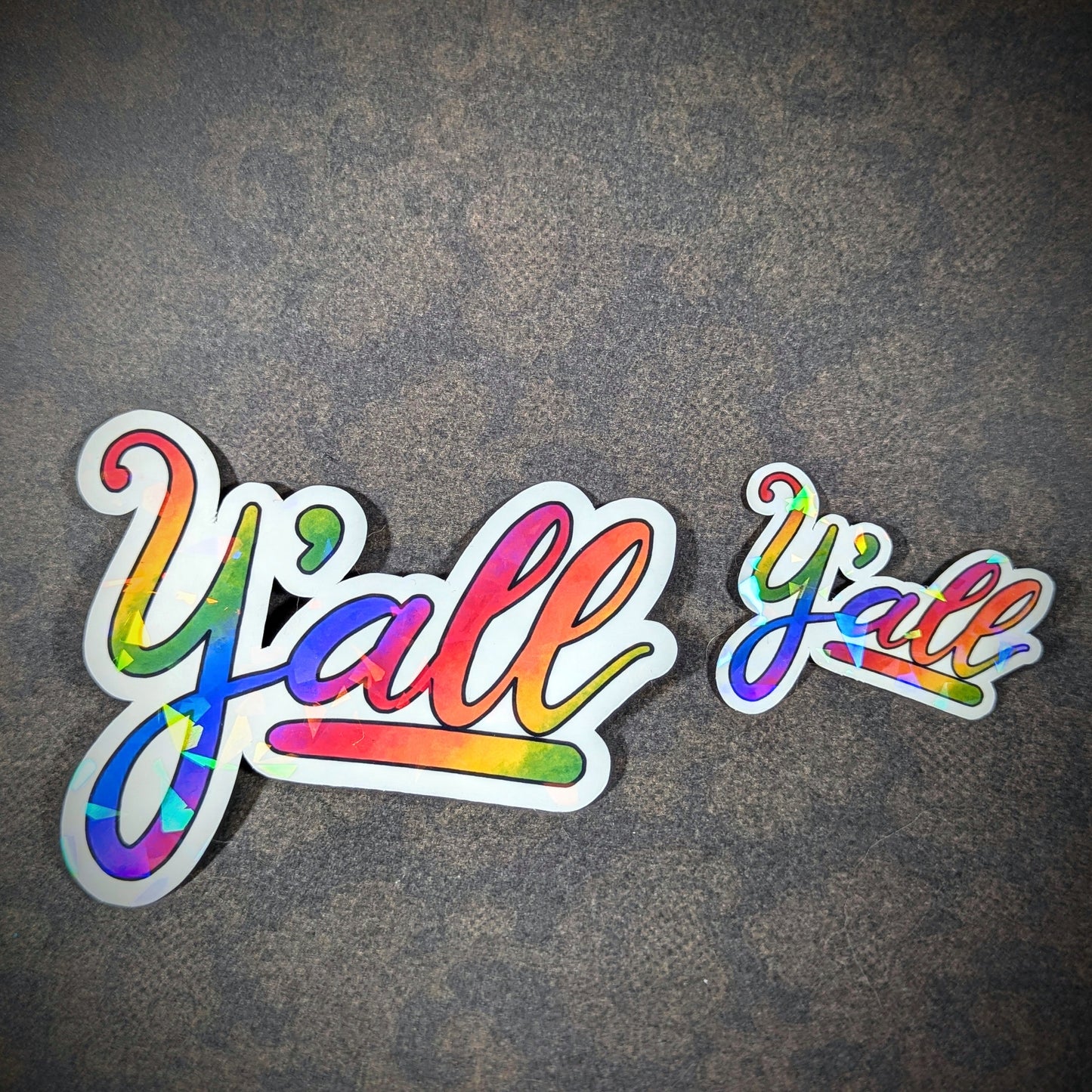 "Large and small colorful rainbow 'Y'all' stickers with holographic shine, featuring a vibrant design perfect for adding Midwestern, Southern charm and LGBTQ+ pride to laptops, notebooks, and personal items."