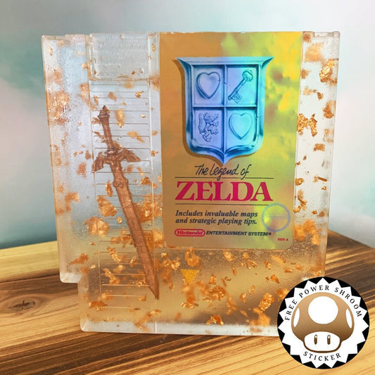 Legend of Zelda resin art featuring a retro NES cartridge with a gold sword design and gold flakes embedded. This handmade display is perfect for gamers and video game collectors, offering a unique décor piece for any gaming setup or retro collection