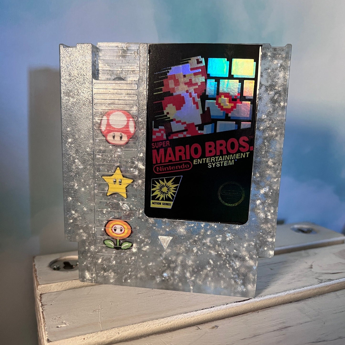 Handmade resin art featuring a retro Super Mario Bros NES cartridge with classic icons like the mushroom, fire flower, and star. Perfect for gamers, collectors, or unique home decor to add a nostalgic touch to any space.