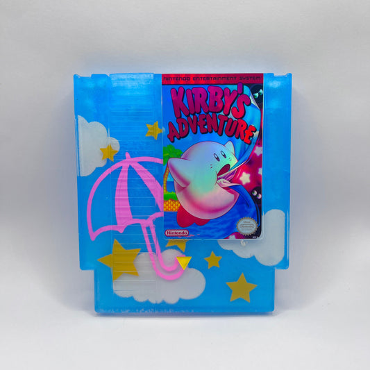 Custom NES cartridge featuring Kirby's Adventure with a colorful design. The blue cartridge has pink umbrellas, clouds, and yellow stars, adding a playful touch for retro gaming fans and collectors. Perfect for showcasing classic Nintendo games.