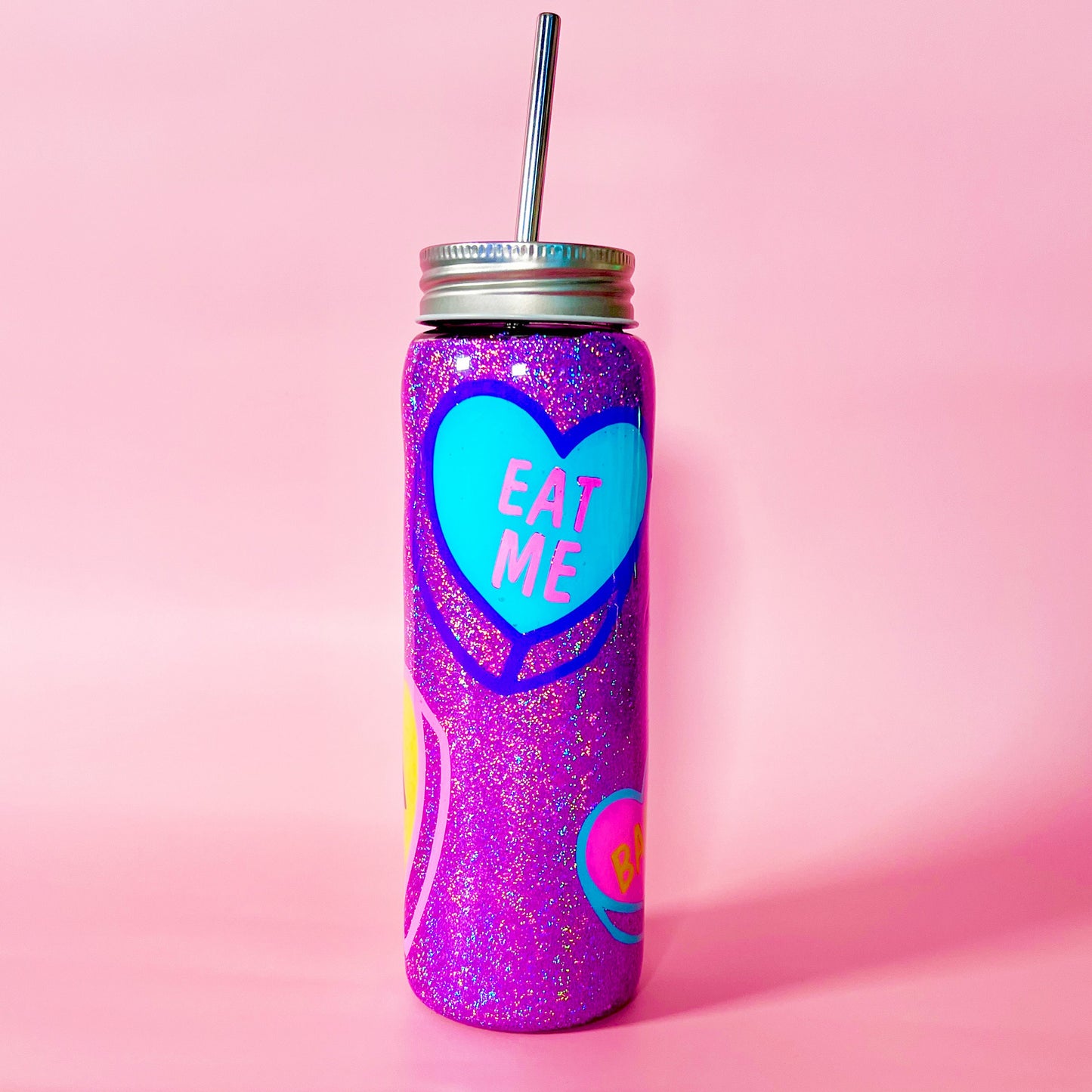 Handmade glitter tumbler with funny candy heart design in vibrant pink, blue, and purple colors. The tumbler features a shiny, sparkly finish and comes with a reusable metal straw. Perfect for adding a fun, quirky touch to your drinkware collection.