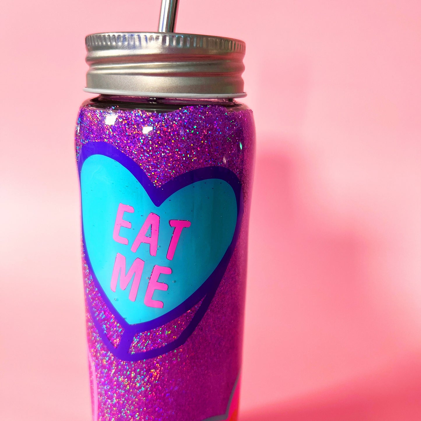 Handmade glitter tumbler with funny candy heart design in vibrant pink, blue, and purple colors. The tumbler features a shiny, sparkly finish and comes with a reusable metal straw. Perfect for adding a fun, quirky touch to your drinkware collection.