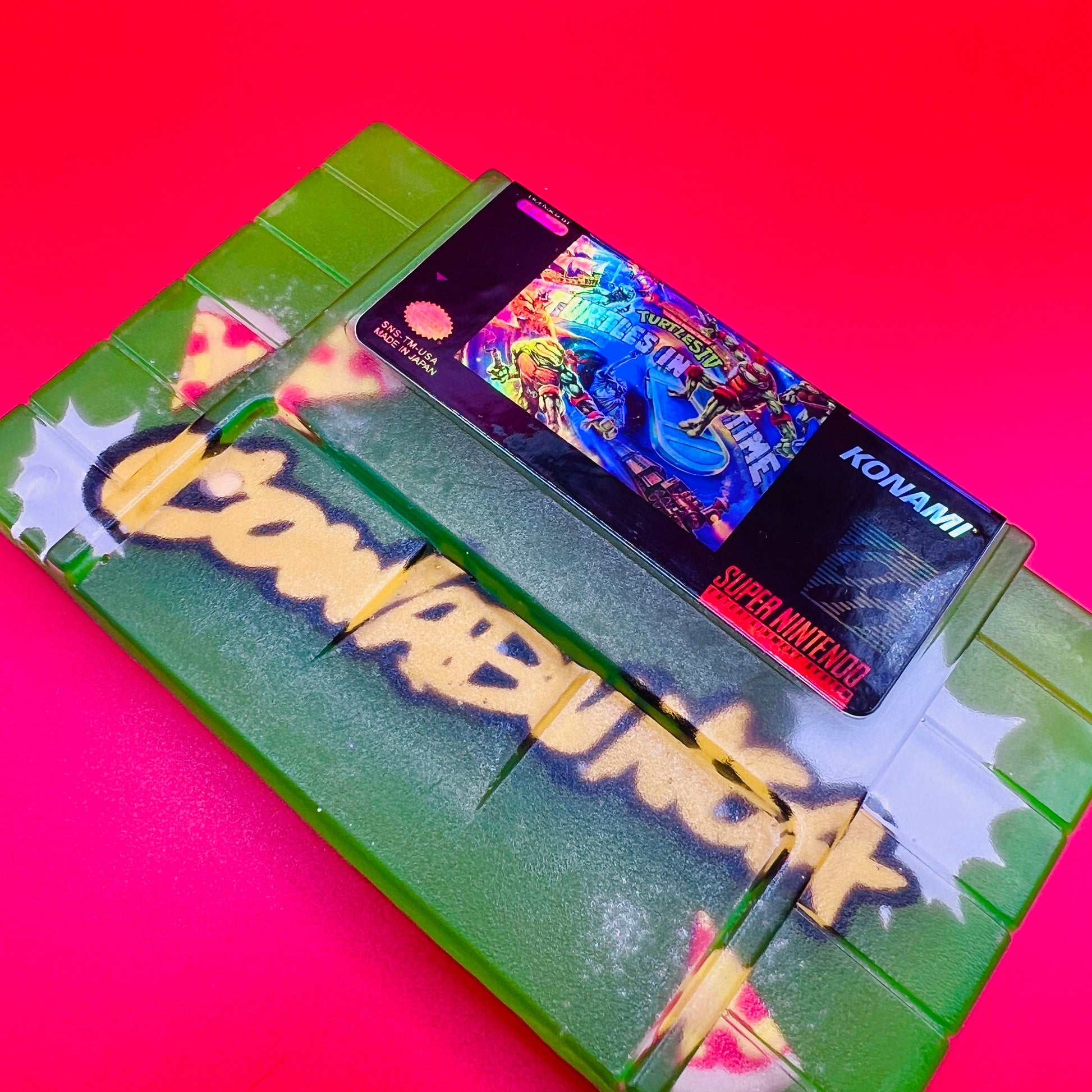 Handmade resin replica of the classic SNES game Turtles in Time. Vibrant green design with graffiti-style elements and retro cartridge art. Perfect for gamers, collectors, or as a unique shelf display for TMNT fans