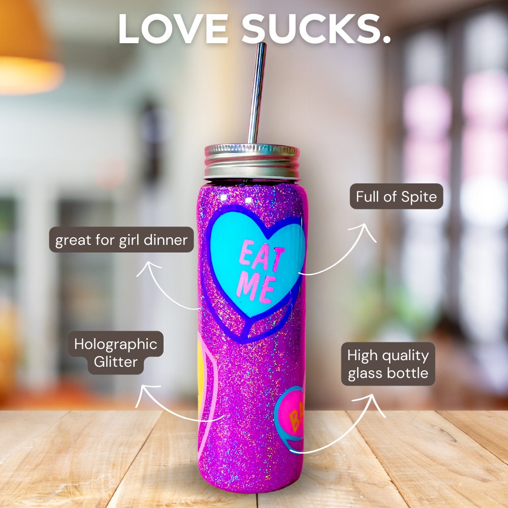 Handmade glitter tumbler with funny candy heart design in vibrant pink, blue, and purple colors. The tumbler features a shiny, sparkly finish and comes with a reusable metal straw. Perfect for adding a fun, quirky touch to your drinkware collection.