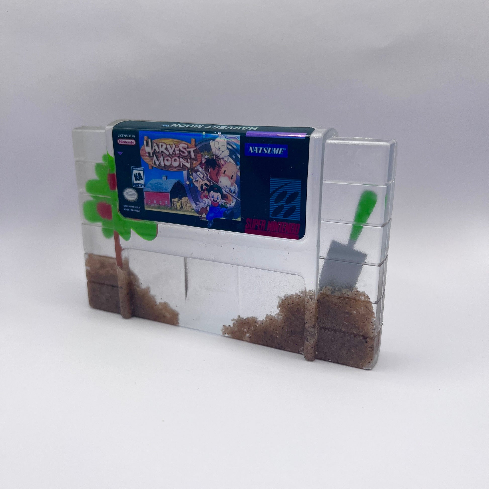 Custom resin art featuring a clear Super Nintendo game cartridge of Harvest Moon filled with decorative sand and tiny plant accents. Perfect as a nostalgic, geek-inspired desk or shelf display for retro gaming enthusiasts.