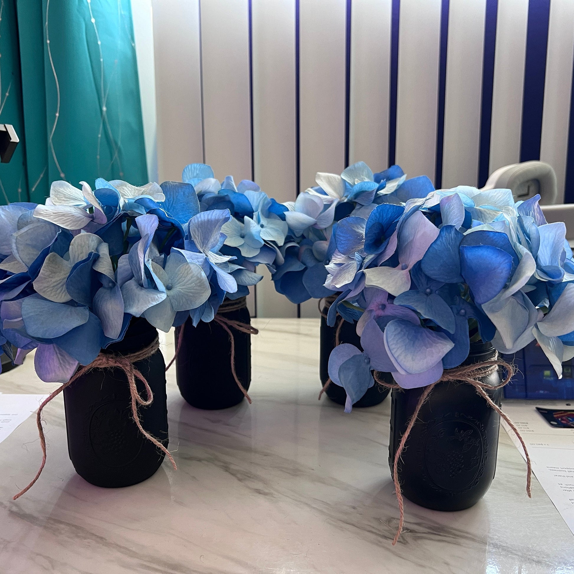 Hand-painted black mason jar with faux blue hydrangea flowers tied with rustic twine. This farmhouse-inspired vase adds a touch of cozy charm to any space, perfect for home decor or special events.