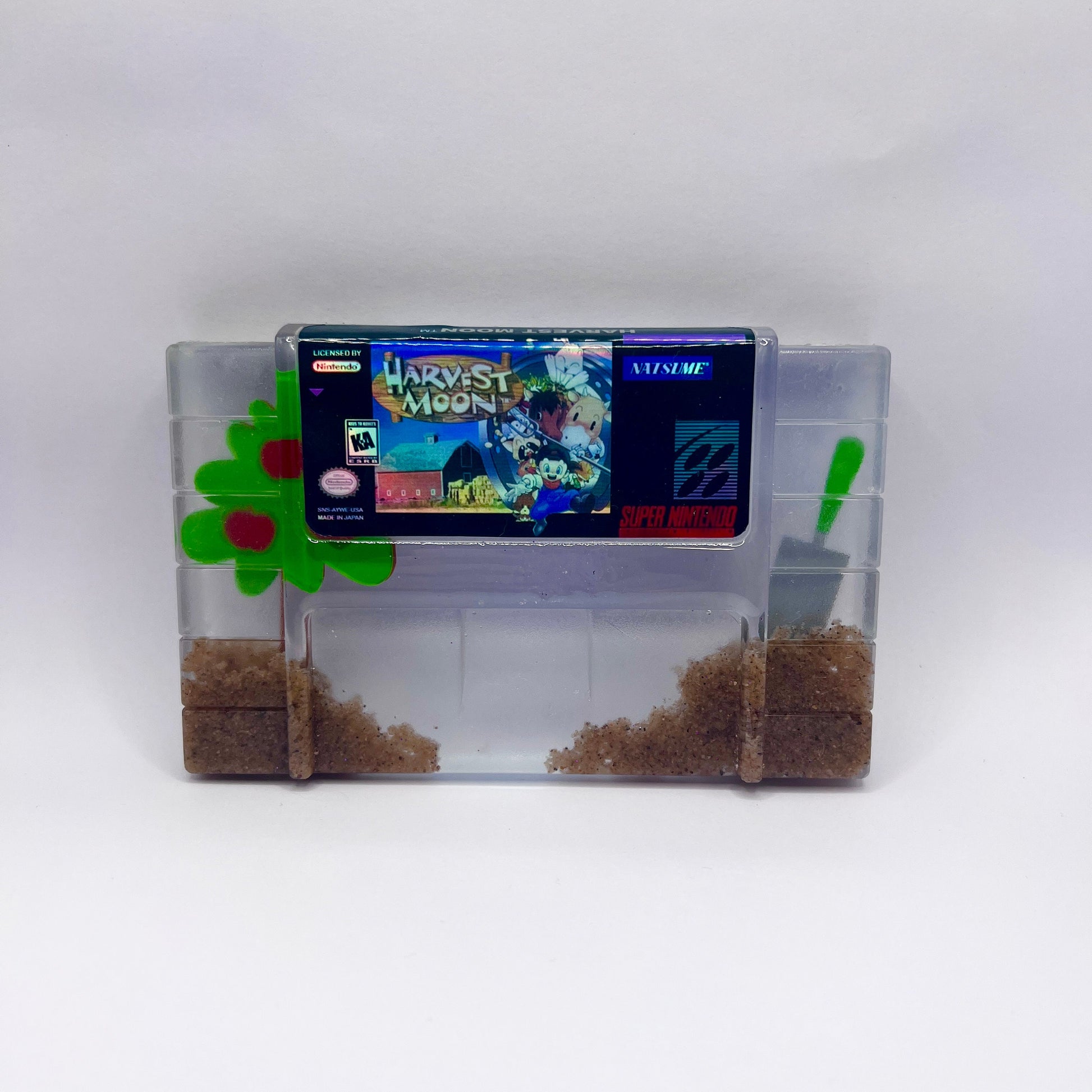 Custom resin art featuring a clear Super Nintendo game cartridge of Harvest Moon filled with decorative sand and tiny plant accents. Perfect as a nostalgic, geek-inspired desk or shelf display for retro gaming enthusiasts.