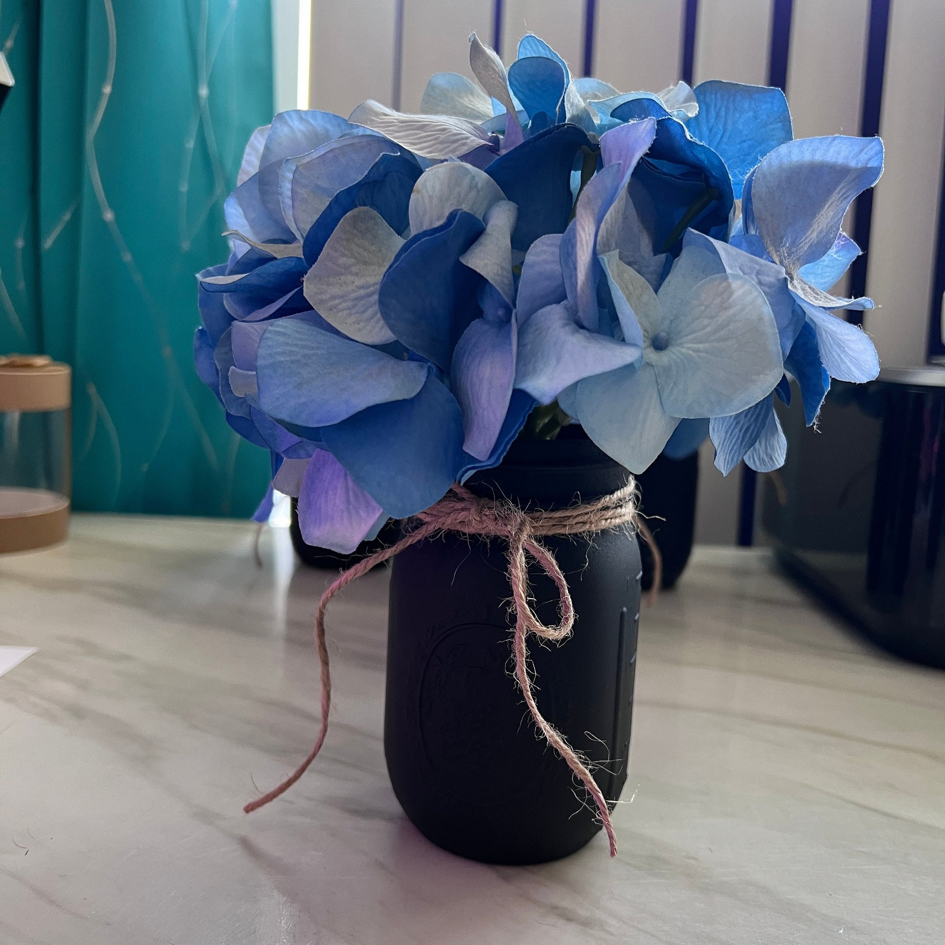 Hand-painted black mason jar with faux blue hydrangea flowers tied with rustic twine. This farmhouse-inspired vase adds a touch of cozy charm to any space, perfect for home decor or special events.