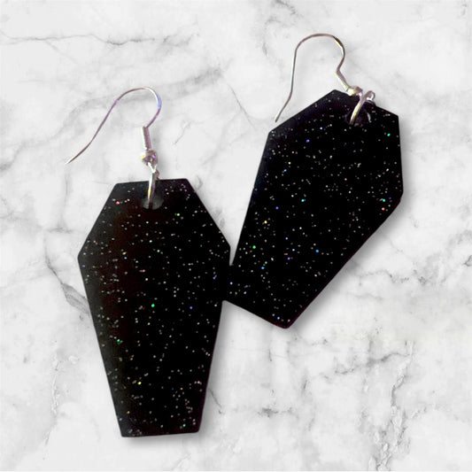 Black holographic glitter coffin-shaped earrings with silver hooks, perfect for gothic and alternative fashion. These statement earrings feature a sparkle finish, adding a spooky yet stylish touch to any outfit. Ideal for Halloween or everyday wear.