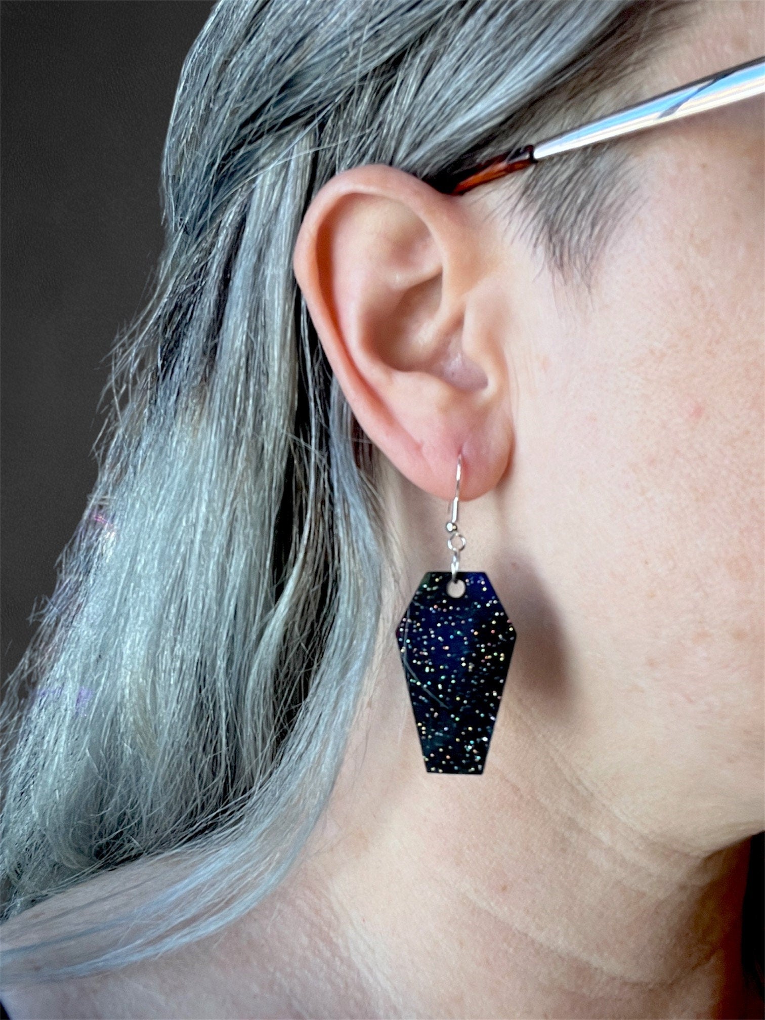 Black holographic glitter coffin-shaped earrings with silver hooks, perfect for gothic and alternative fashion. These statement earrings feature a sparkle finish, adding a spooky yet stylish touch to any outfit. Ideal for Halloween or everyday wear.