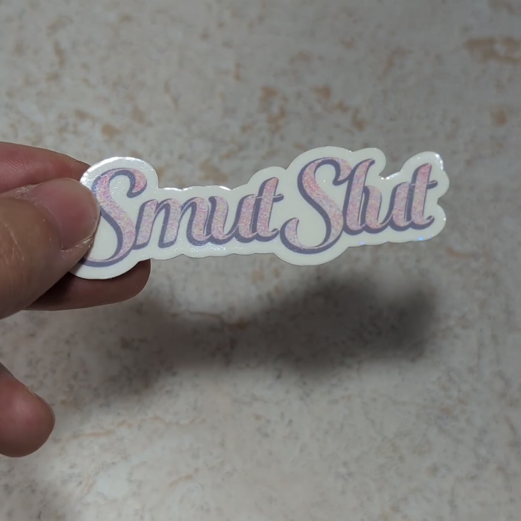 Video of a hand holding "Smut Slut" sticker with elegant pink speckled text, to demonstrate holographic properties, perfect for book lovers and spice enthusiasts. Set against a cozy book stack background, this design adds a bold, fun flair to your aesthetic.