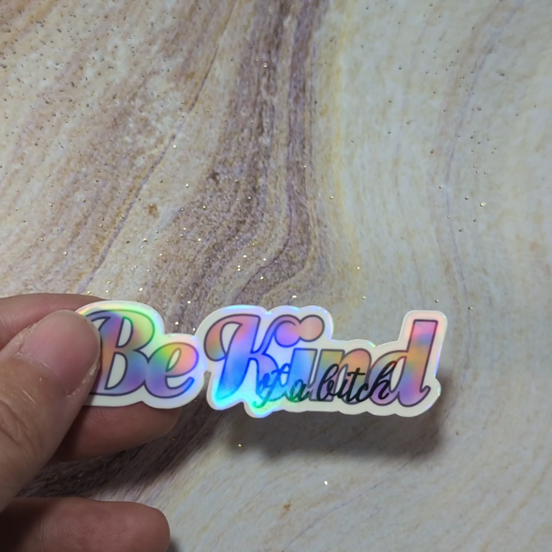 Video of a hand holding "Be Kind of a Bitch" holographic sticker with bold rainbow text, perfect for empowering vibes and showcasing personality. Set on a textured background, this vibrant design is edgy and inspiring.