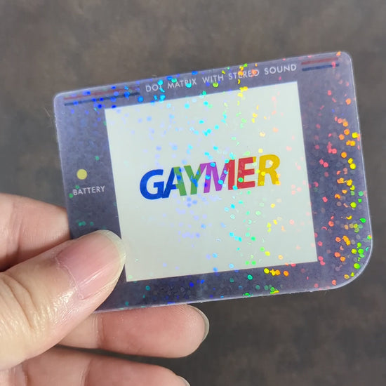 "Video of hand holding sticker to show holographic effect. Holographic sticker with 'Gaymer' in rainbow text on Game Boy screen, inspired by classic Game Boy design with 'Dot Matrix with Stereo Sound' label. A playful, pride-themed decor piece for LGBTQ+ gamers."