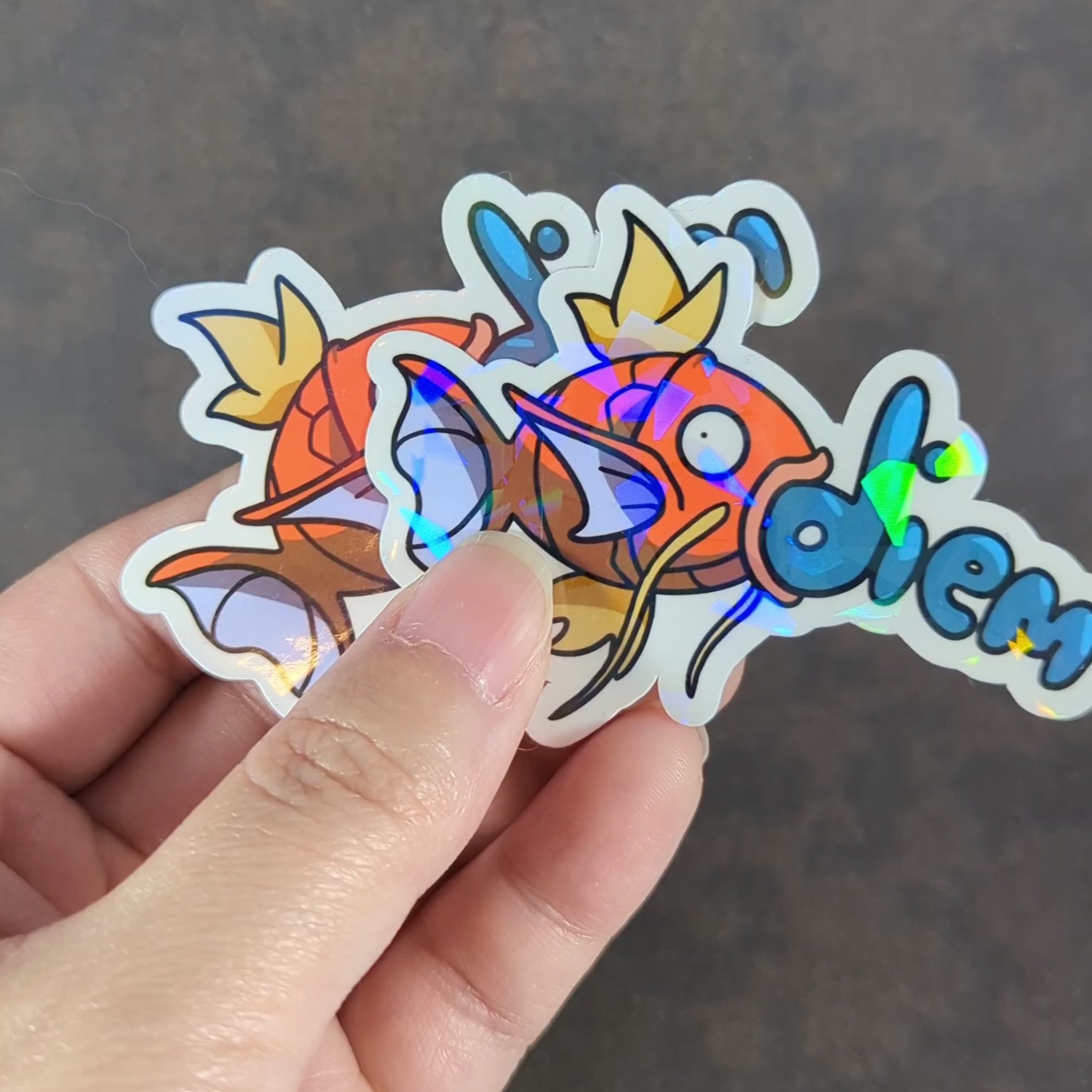 "Video of plain and Holographic 'Carpe Diem' inspired stickers featuring a vibrant Magikarp Pokemon design, perfect for water bottles, laptops, and notebooks. Adds a fun, motivational touch for gamers and fish enthusiasts alike."