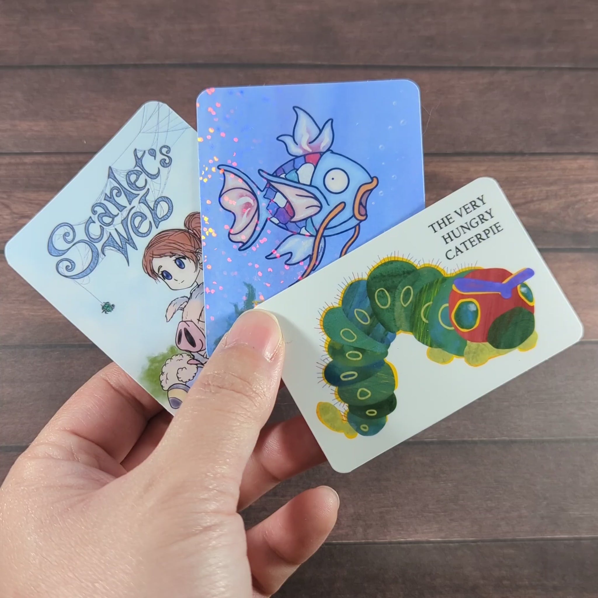 
"Video of a hand holding vinyl stickers to show off glossy and holographic finishes, inspired by storybook themes: Scarlet's Web, The Rainbow Karp, and The Very Hungry Caterpie, designed for collectors and gamers, displayed on a wooden background."