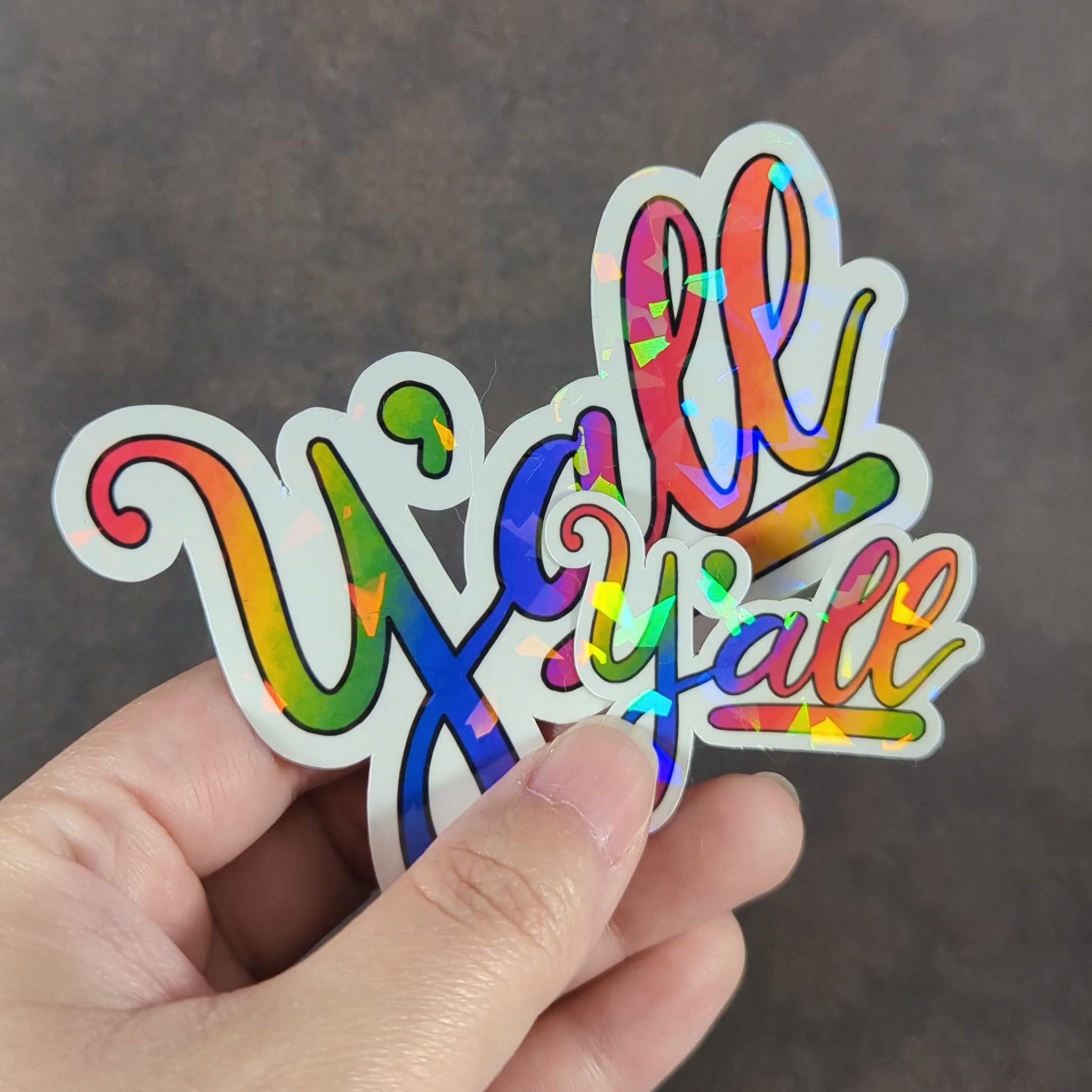 "Video showing holographic effect of Large and small colorful rainbow 'Y'all' stickers with holographic shine, featuring a vibrant design perfect for adding Midwestern, Southern charm and LGBTQ+ pride to laptops, notebooks, and personal items."